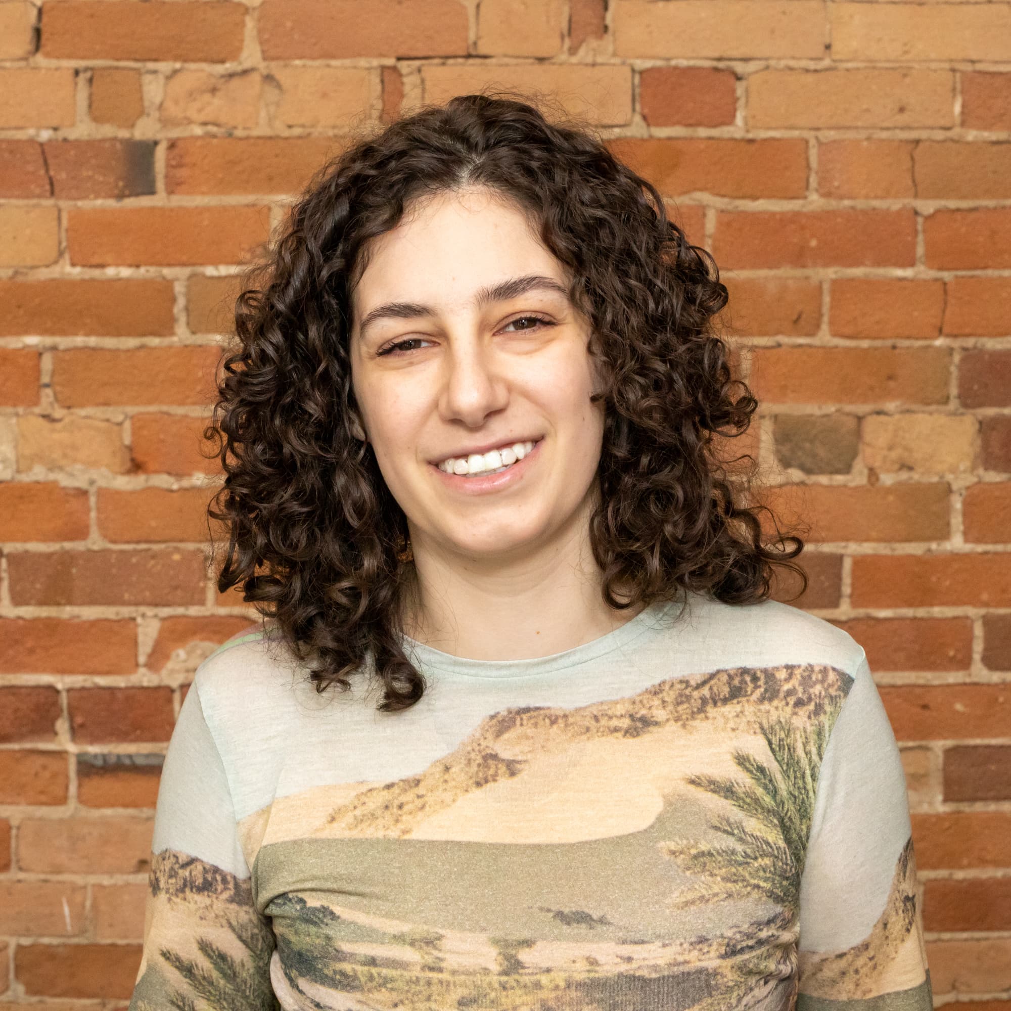 Meredith has curly, shoulder-length hair and bangs framing their face as they look at the camera smiling